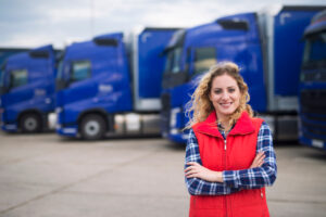 A blonde, female truck driver stands in front of a row of semi trucks, she is wearing a blue flannel with a red puffer vest, with her arms crossed, she's smiling knowing that she has solid data and information to increase the kpi for trucking companies