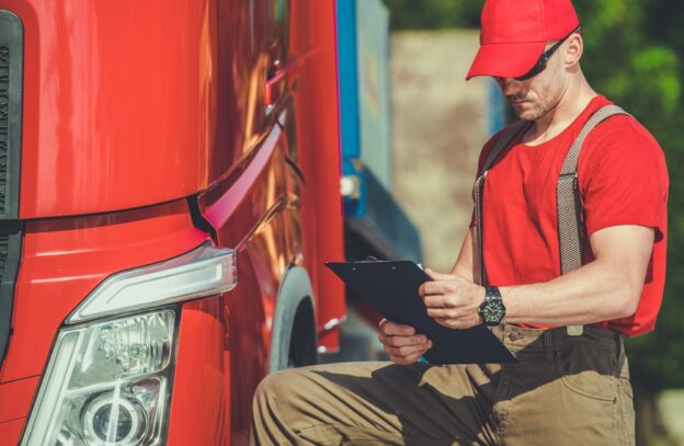 A semi truck driver in a red shirt and cap reviews inspection paperwork while crouching next to a large red truck. The sun is shining, highlighting the busy work environment